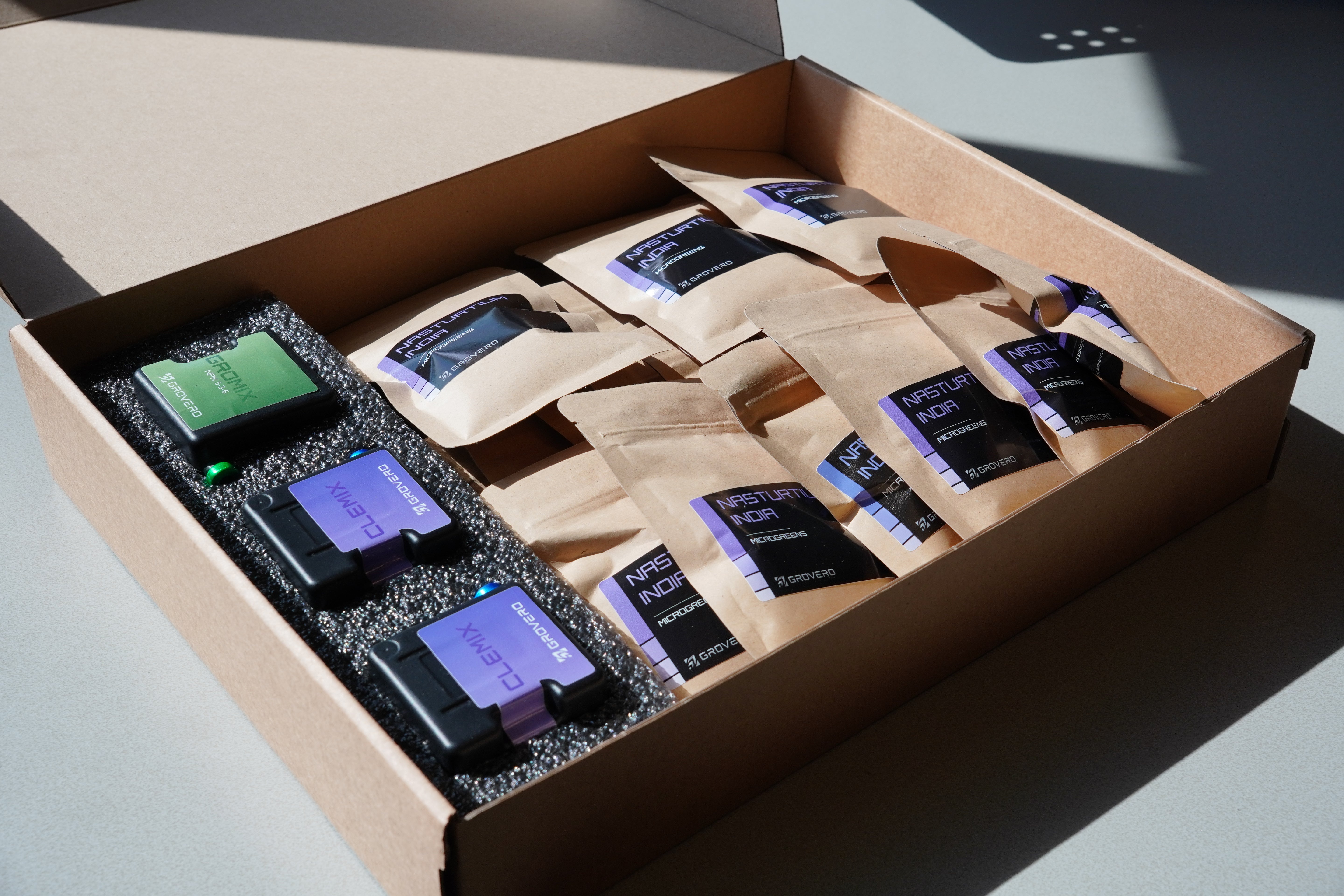 An open kraft cardboard box containing multiple small brown paper pouches and two black rectangular cartridges with purple and green labels, photographed on a light surface in bright natural light.