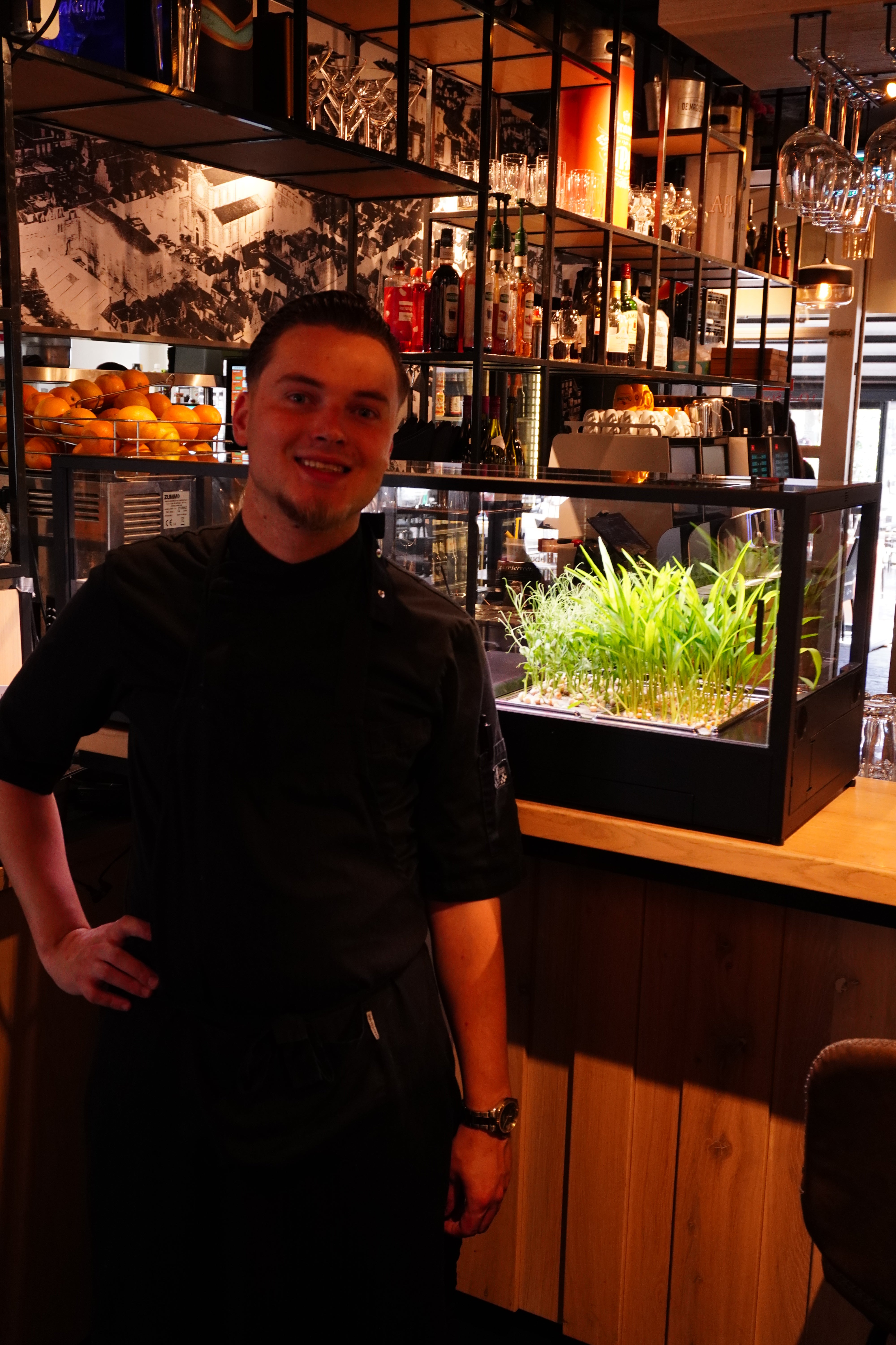 Man standing in a bar setting with shelves of drinks and an indoor microfarm.