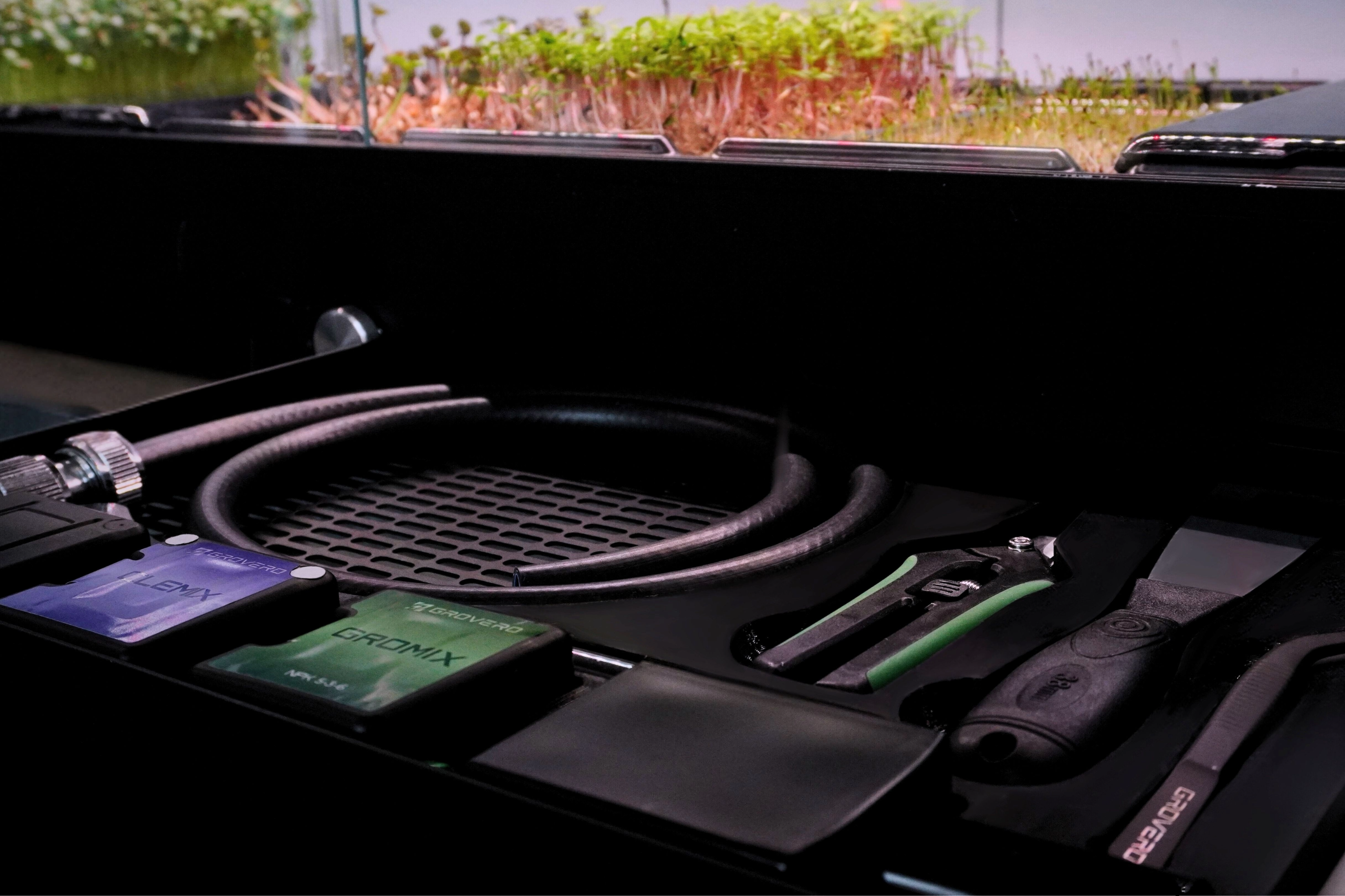 A black grow kit organizer tray containing a coiled black hose and hand tools, photographed in a dim indoor setting with plants visible in the background.