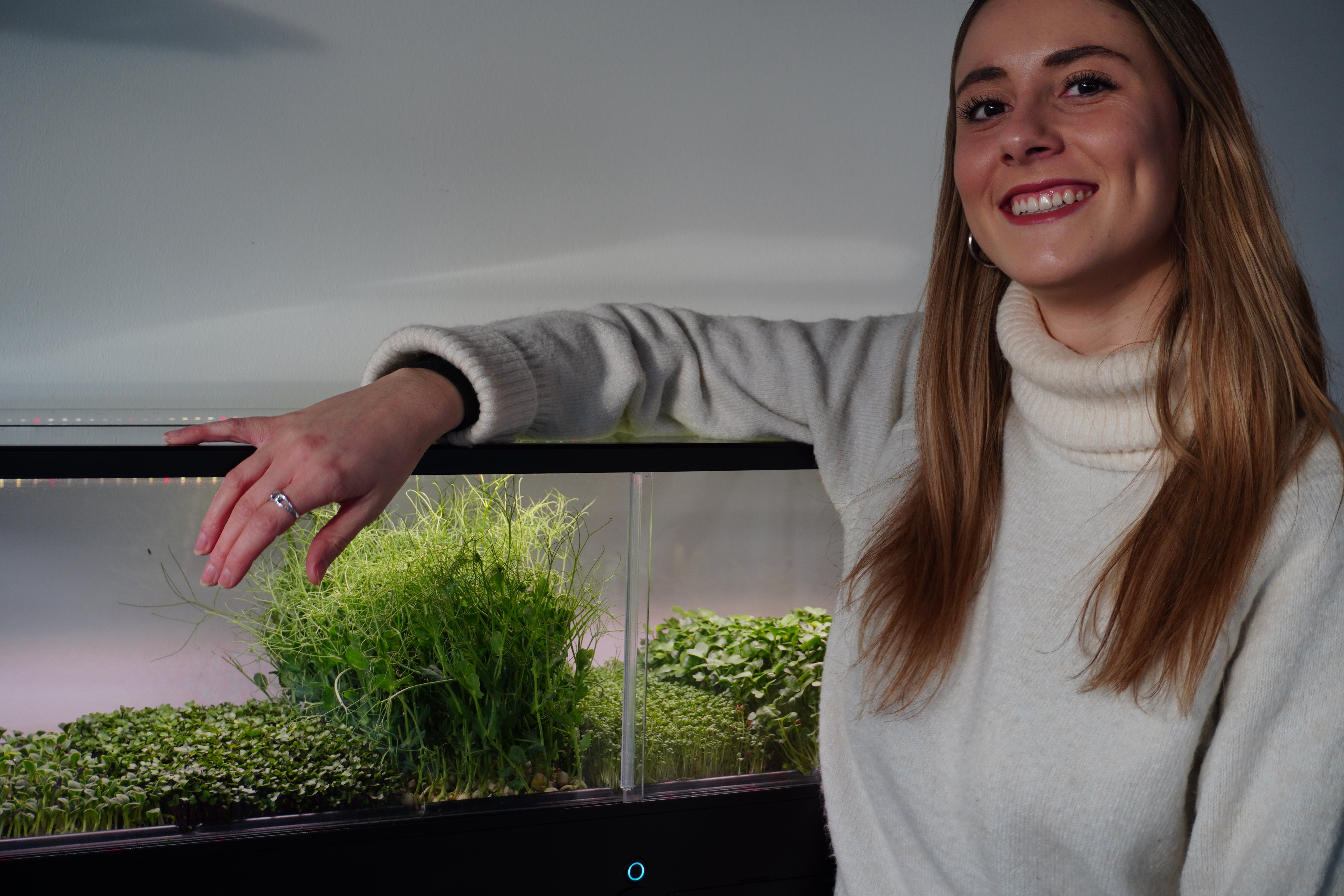 A smiling woman with long light brown hair leans her arm over a modern indoor microfarm filled with green microgreens and herbs under grow lights. She is wearing a light gray turtleneck sweater.