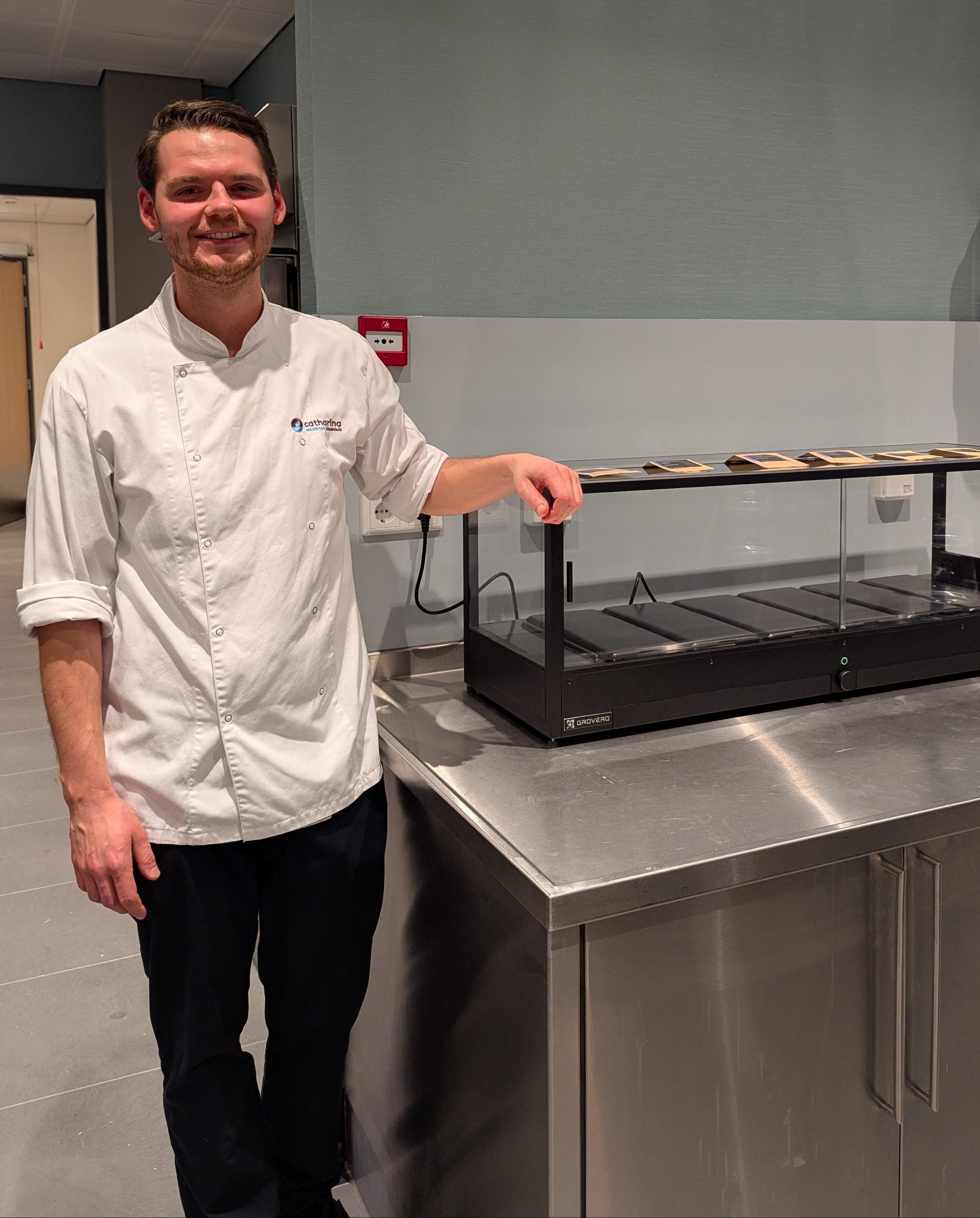 A man in a white chef's coat stands next to a black microfarm on a stainless steel counter in a kitchen setting.
