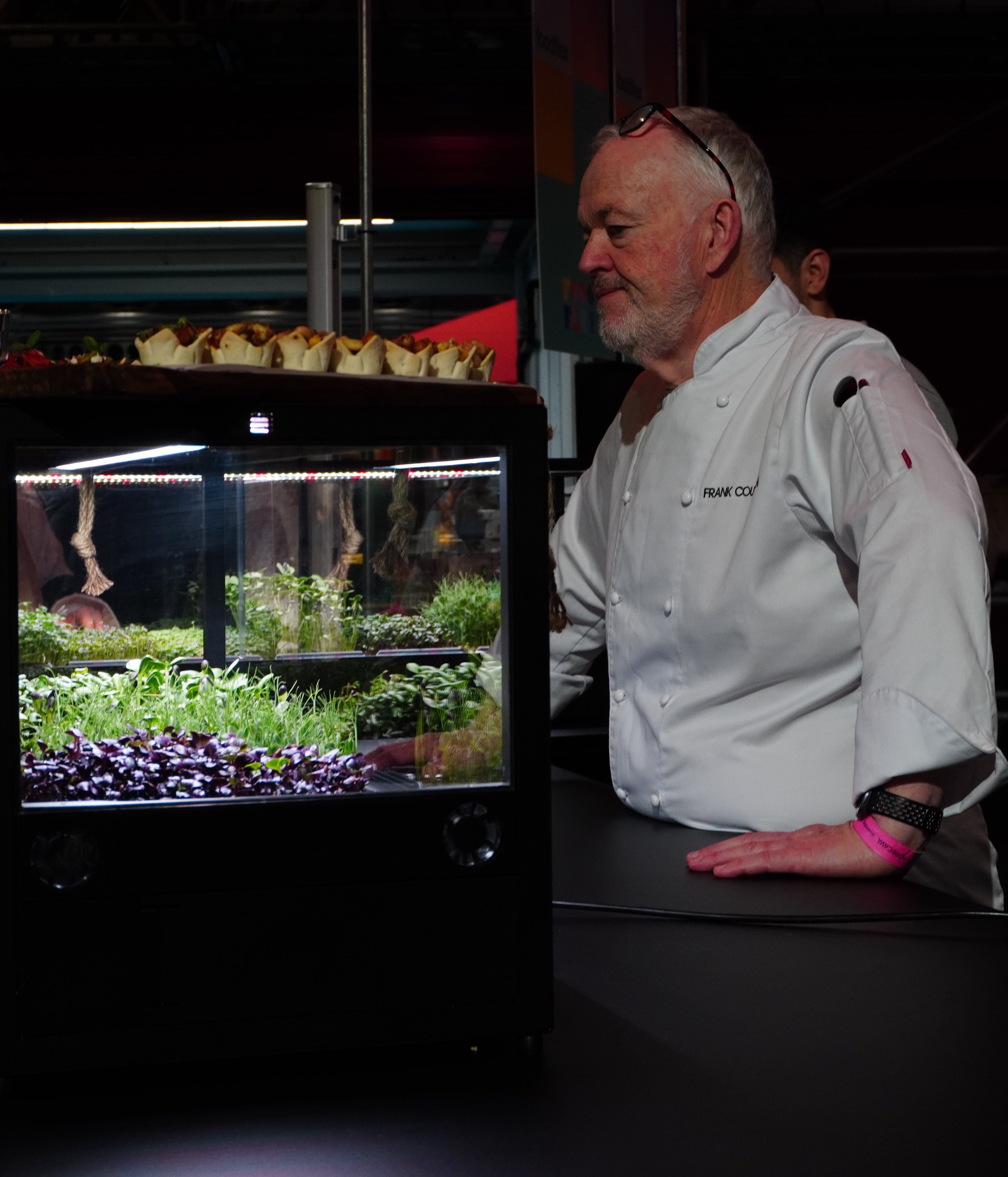 A chef in a white uniform stands beside a countertop hydroponic microfarm showing green plants growing under LED lighting.