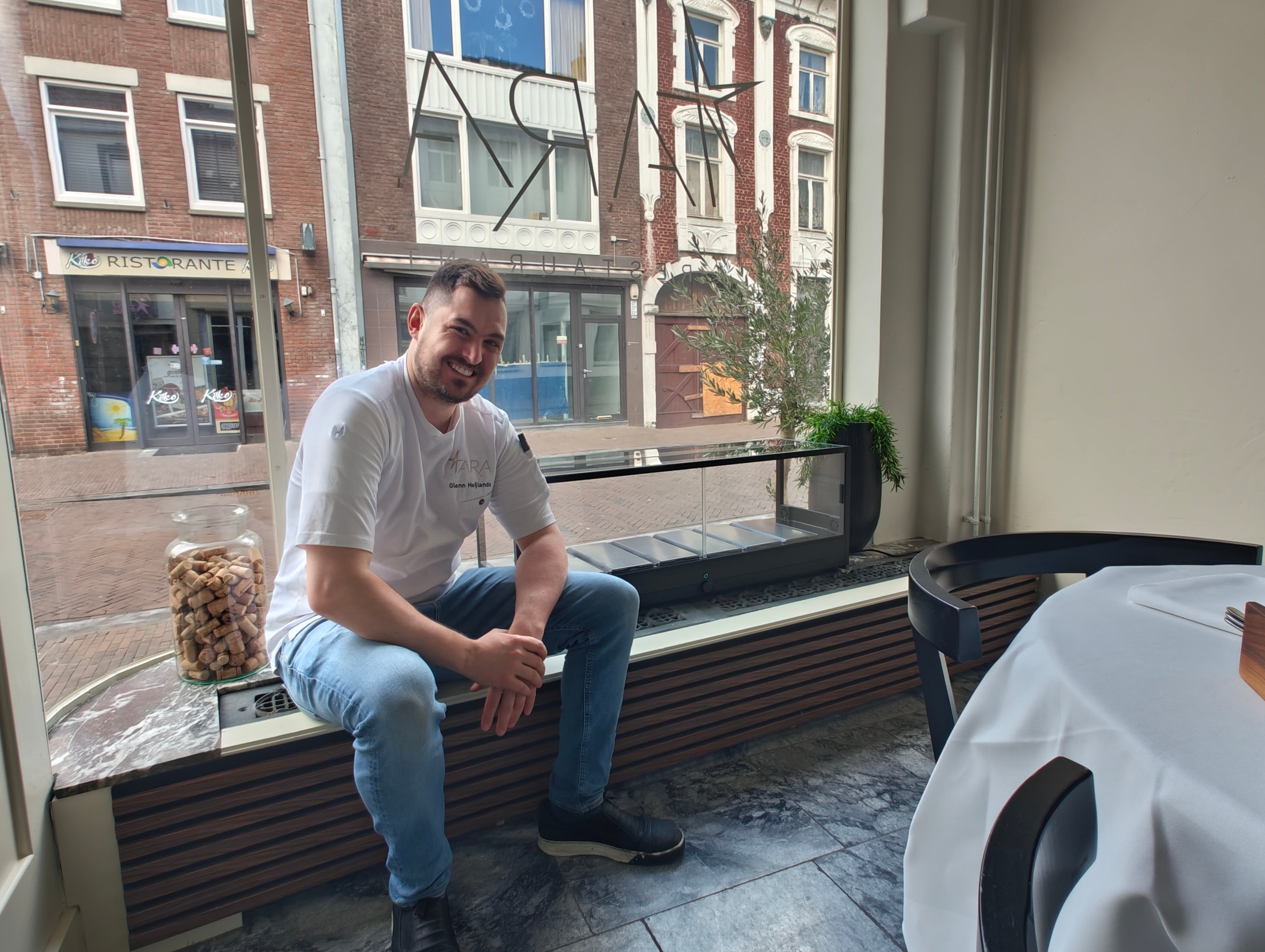A man wearing a white t-shirt and blue jeans sits smiling on a window ledge inside a restaurant, with a street scene visible outside and an indoor microfarm sitting next to him.