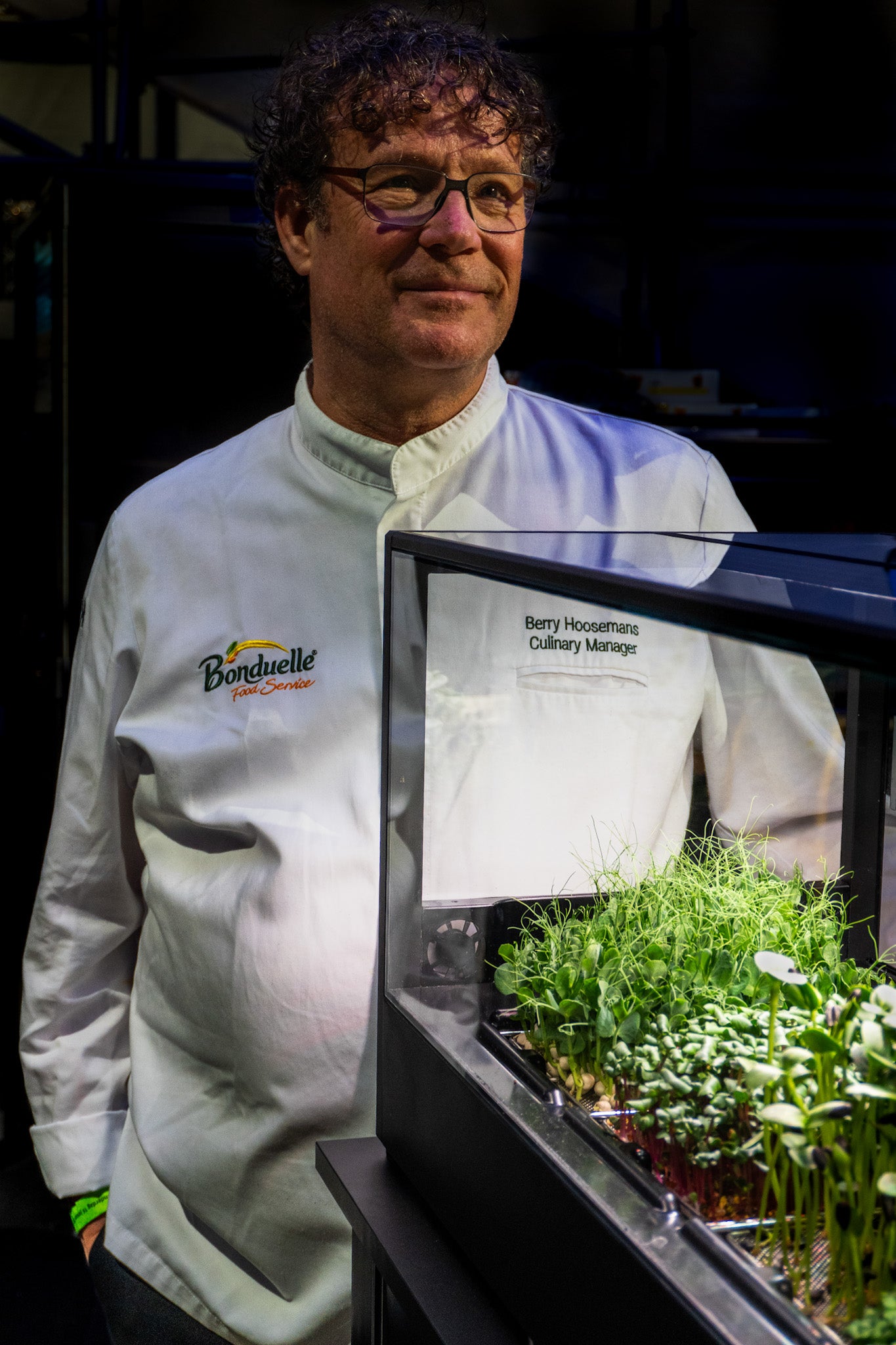 Person wearing a chef's coat with a brand logo, standing next to a small indoor microfarm.